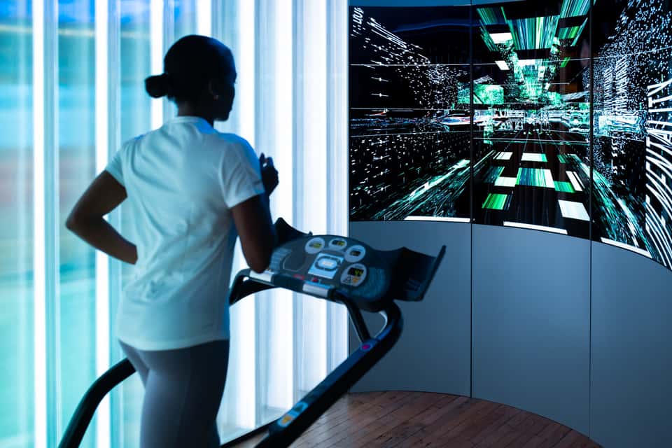 Photo of Adidas Running Lab installation in London showing a runner on a treadmill in front of a large LED screen displaying an abstract visualization of a cityscape.