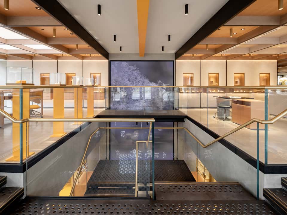 Inside the VASHI store, a central staircase with glass railings, wood accents, and a large digital screen on the far wall. Desks and shelves are visible in the background.